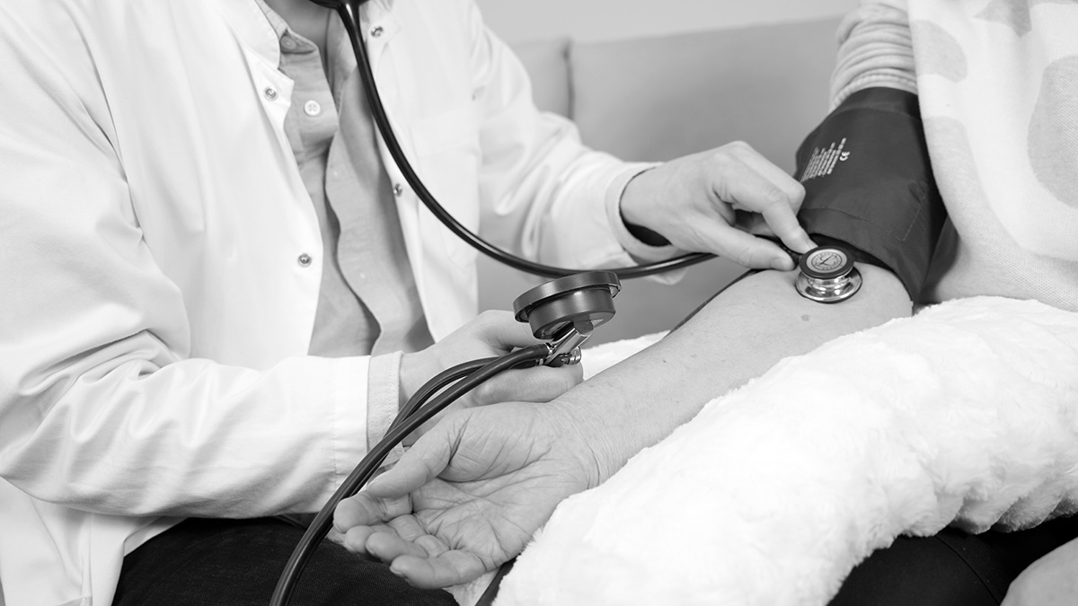 Schwarzweißfoto: Ärztin misst mit Blutdruckmanschette und Stethoskop den Blutdruck am Oberarm einer Patientin.