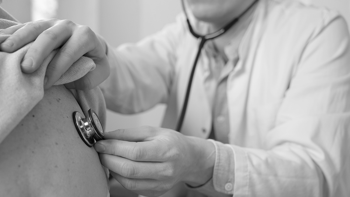 Schwarzweißfoto: Arzt hört mit dem Stethoskop die Lunge eines Patienten am Rücken ab.